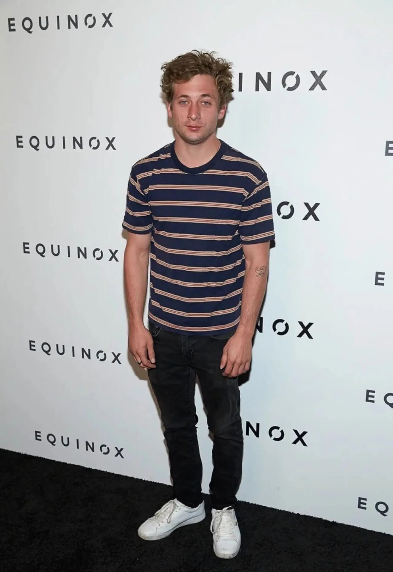 Jeremy Allen White Awkward meme template - Jeremy Allen White stands on a black carpet at an Equinox event, navy striped t-shirt, dark jeans, white sneakers. His arms h